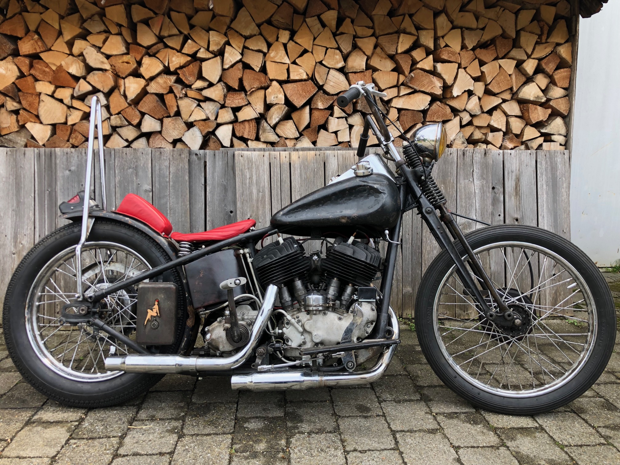 Old-School-Chopper mit mattschwarzem Tank, roter Sitzbank und Sissybar vor einem Holzstapel