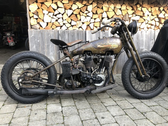 Bobber mit rostbrauner Tankpatina und Harley-Schriftzug, aufgebockt vor dem Holzstapel