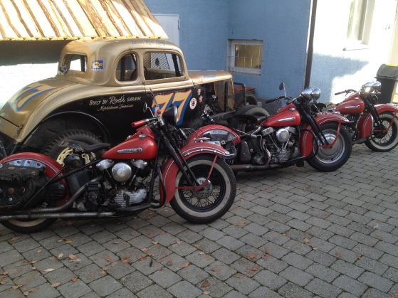 Zwei rote Harleys vor einem goldenen Hot-Rod-Coupé auf dem Pflasterhof