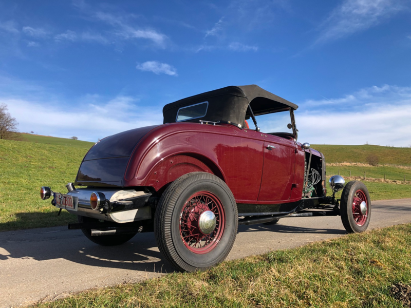 Weinroter Roadster mit roten Speichenfelgen auf einer Landstraße vor grünen Hügeln