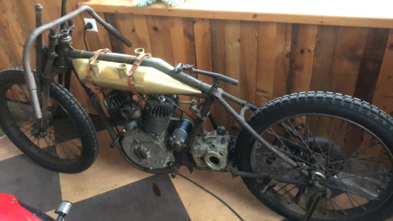 Angefangenes Projekt aus nacktem Rahmen, Motor und Messingtank, festgezurrt mit Lederriemen